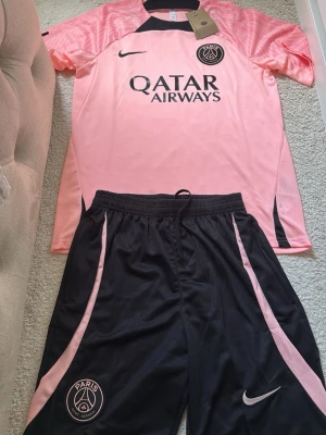 PSG matchställ rosa/svart Nike - Säljer ett fett PSG matchställ från Nike med rosa tröja och svarta shorts med rosa detaljer. Tröjan har Qatar Airways-tryck och PSG-logga på bröstet, shortsen har PSG-märke och Nike swoosh. Perfekt för fotbollsträning eller att chilla i. Materialet är lätt och andas.