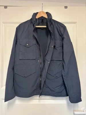 Peak Performance, tunn, mörkblå, strl M, fältjacka, nyskick, bomull/nylon - Säljer en tunn mörkblå jacka i storlek medium från Peak Performance, modell Hunt J, M65-jacka/fältjacka/militärjacka, i nyskick. Nypris var över 2000 kr. Perfekt för sommaren, även sen vår och tidig höst. Dold huva, dragsko i midjan och många fickor.   Bröstmått (armhåla till armhåla): C:a 110 cm. Rygglängd: 72 cm.   Kontakta mig gärna vid frågor.