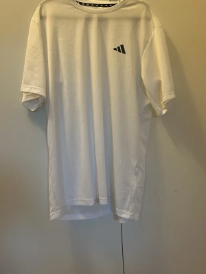 Vit t-shirt från Adidas - Vit t-shirt från Adidas med svart logga på bröstet. Klassisk passform med korta ärmar och rund halsringning. Tillverkad i ett lätt och ventilerande material som passar perfekt till vardags eller träning.