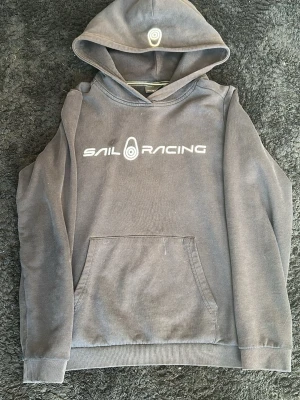 Grå hoodie från Sail Racing - Mörkgrå hoodie från Sail Racing med vit logga och text på bröstet. Klassisk känguruficka framtill och ribbade muddar vid ärmslut och nederkant. Huva med tryckt logga på toppen. Perfekt för en avslappnad och sportig stil.