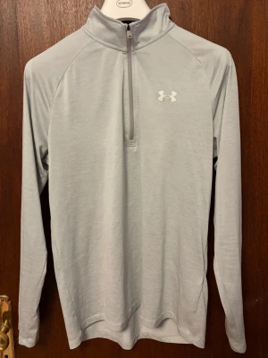 Ljusgrå träningströja från Under Armour - Snygg ljusgrå långärmad träningströja från Under Armour med halv dragkedja framtill och hög krage. Tillverkad i ett lätt och stretchigt funktionsmaterial som passar perfekt för löpning eller gym. Diskret logga på bröstet och nacken.