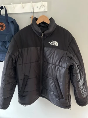 Svart pufferjacka från The North Face - Säljer en svart pufferjacka från The North Face med vit logga på bröstet. Jackan har dragkedja framtill, två sidofickor och justerbar dragsko i nederkant. Tillverkad i slitstarkt syntetmaterial och perfekt för kalla vinterdagar. Den är använd litegrann under en vinter men är nytvättad och i väldigt bra skick! Jackan köptes för 2600kr. Buda gärna 