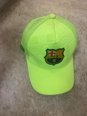Neongul FC Barcelona keps - Snygg neongul keps med FC Barcelona-logga framtill och mesh på sidorna för extra ventilation. Justerbar baktill och broderad text på insidan. Perfekt för dig som vill sticka ut och visa vilket lag du hejar på.