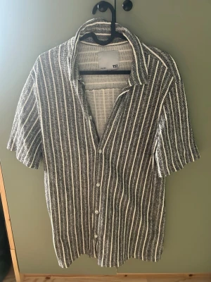 Randig skjorta från 157 med kort ärm - Säljer en snygg skjorta från 157 med vit och grå randig design. Skjortan har korta ärmar, klassisk krage och knappar framtill. Perfekt för dig som gillar en avslappnad men stilren look.