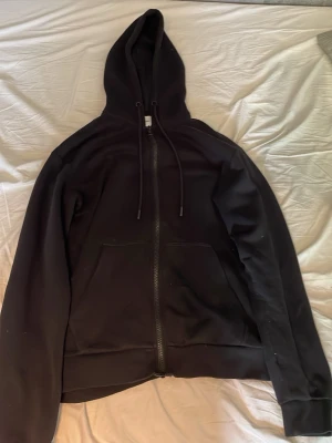 Svart hoodie med dragkedja - En stilren svart hoodie med dragkedja framtill och två stora fickor. Tröjan har huva med dragsko och ribbade muddar vid ärmslut och nederkant. Perfekt för en avslappnad look och enkel att matcha med andra plagg. Tveka inte höra av dig vid minsta fundering ✅🤝