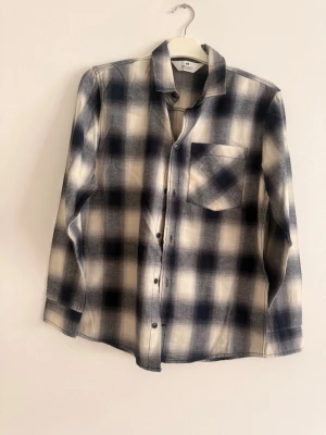 Rutig långärmad skjorta från H&M - Snygg rutig skjorta från H&M i svart, beige och vitt. Skjortan har lång ärm, klassisk krage, bröstficka och knappar framtill. Perfekt för en avslappnad och trendig look.