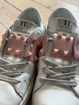 Valentino Rockstud Untitled sneakers - Snygga vita sneakers från Valentino med ljusrosa detaljer och ikoniska nitar på sidorna och ovandelen. Skorna har vita skosnören och en klassisk låg siluett. Tillverkade i läder med gummisula och coola detaljer som sticker ut.