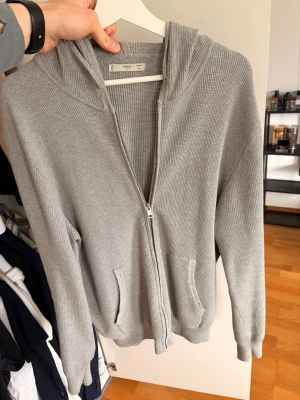 Ljusgrå ribbad hoodie från MNG - Ljusgrå hoodie med dragkedja från ARKET. Tillverkad i mjukt ribbat material, har två fickor framtill och en skön huva. Perfekt för en chill och avslappnad stil. Passar till det mesta och är enkel att matcha.