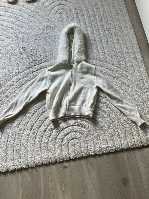 Vit hoodie med fuskpäls luva - Mysig vit hoodie med dragkedja och fluffig fuskpäls runt huvan. Tröjan har ribbade muddar vid ärmslut och nederkant samt en croppad passform. Pälsen går även att ta av o på med hjälp av knappar och dragkedja går att justera från båda hållen. Säljer för den inte passar