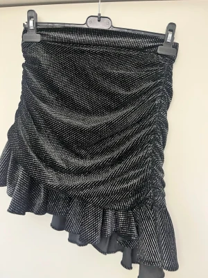 Glittrig svart kortkjol med volang - Supersnygg svart kortkjol med glittriga detaljer och volangkant. Kjolen har en draperad, rynkad design som ger en cool och trendig look. Perfekt för dig som vill sticka ut! Asymmetrisk nederkant och stretchigt material för skön passform. Oanvänd 