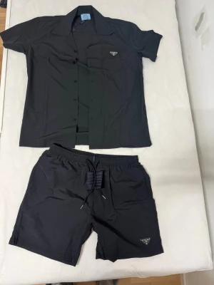 Svart set från Prada med skjorta & shorts - Snyggt svart set från Prada med kortärmad skjorta och matchande shorts. Båda plaggen har diskret Prada-logga framtill. Skjortan har knappar och klassisk krage, shortsen har resår och dragsko i midjan. Perfekt för en trendig och avslappnad look.
