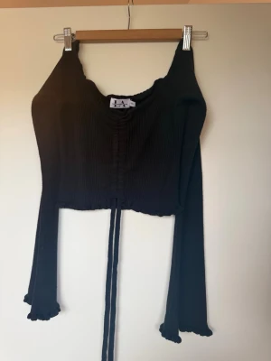 Svart croppad topp från NA-KD - Trendig svart croppad topp från NA-KD med långa trumpetärmar och rynkdetalj framtill. Tillverkad i ribbat stretchigt material och har knytband i midjan. Perfekt för dig som gillar en edgy och modern stil.