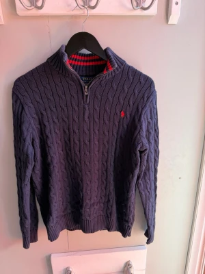 Mörkblå kabelstickad tröja Polo Ralph Lauren - Kabelstickad mörkblå tröja från Polo Ralph Lauren med halv dragkedja och röd broderad logga på bröstet. Tröjan har ribbade muddar och krage samt insida i rött och marinblått randigt mönster. Perfekt för lager på lager och klassisk stil.