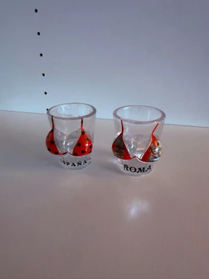 Shotglas souvenir Spanien & Rome - Två unika shotglas i glas med 3D-design av röda bikinis på framsidan. Ena glaset har texten 'ESPAÑA' och svarta prickar, det andra 'ROMA' med motiv från Rom. Perfekta som roliga samlarprylar eller partyaccessoarer. 40kr för båda, 20kr st