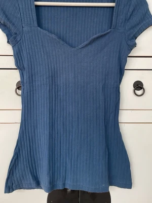 Blå ribbad topp med v-ringning - Snygg blå ribbad topp med v-ringad hals och korta ärmar. Toppen har en figurnära passform och är tillverkad i mjukt bomullsmaterial. Perfekt att matcha med jeans eller kjol för en enkel och stilren look. Storlek S