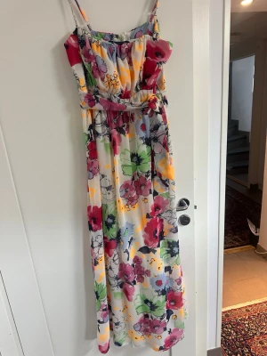 Blommig långklänning med smala axelband - Färgglad långklänning med blommigt mönster i rött, grönt, lila och gult. Klänningen har smala axelband och ett band i midjan för en feminin siluett. Perfekt för varma dagar och somriga tillfällen.