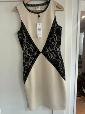 Beige klänning med svart spets - Snygg beige kortklänning från Molly Bracken med svarta spetsdetaljer i sidorna och runt halsen. Ärmlös modell med figurnära passform och elegant spetsmönster som ger en lyxig känsla. Perfekt för dig som vill sticka ut.