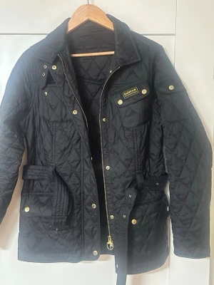 Svart quiltad vindjacka från Barbour - Snygg svart quiltad vindjacka från Barbour med guldiga knappar och dragkedja. Jackan har bälte i midjan, flera fickor och en klassisk Barbour-logga på bröstet. Perfekt för dig som vill ha en stilren och tidlös jacka med coola detaljer.