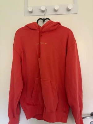 Röd/orange hoodie från Adidas - Säljer en röd/orange hoodie från Adidas med liten logga på bröstet. Hoodien har huva med dragsko och en stor magficka framtill. Tillverkad i mjukt bomullsmaterial och har ribbade muddar vid ärmslut och nederkant. Perfekt för en avslappnad och sportig stil.