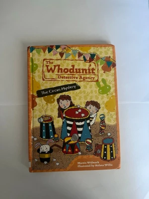The Whodunit Detective Agency - En färgglad och illustrerad bok på engelska, 'The Whodunit Detective Agency: The Circus Mystery', skriven av Martin Widmark och illustrerad av Helena Willis. Omslaget har cirkustema med barn och djur, hårda pärmar och lekfull design.