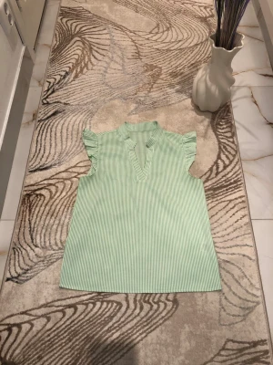 Grönrandig blus med volangärm - Snygg blus med tunna gröna och vita ränder, volangdetaljer vid ärmarna och en liten ståkrage med v-ringning. Perfekt till jeans eller kjol för en fräsch look. Materialet känns lätt och luftigt, troligen bomull eller bomullsblandning.