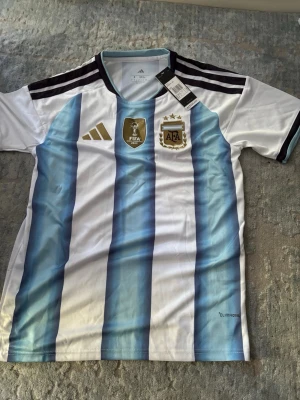 Argentina matchtröja Adidas S - Säljer en officiell Argentina matchtröja från Adidas i storlek S. Tröjan har klassiska blå och vita ränder, svarta detaljer på axlarna och guldiga logotyper på bröstet. Tillverkad i lätt och ventilerande material med Climalite-teknologi.