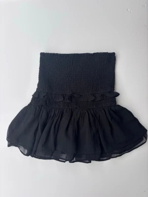 Svart volangkjol från Gina Tricot - Supersöt svart kortkjol från Gina Tricot med smock i midjan och volangdetaljer. Kjolen har en luftig och flowy känsla, perfekt för att styla med en snygg top eller stickad tröja. Snyggt att vika ner kanten lite.  Använd väldigt få gånger.
