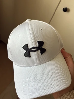 Vit keps från Under Armour - Snygg vit keps från Under Armour med det klassiska svarta logotypen framtill. Kepsen har böjd skärm och är tillverkad i ett slitstarkt, lätt material som andas. Perfekt för dig som vill ha en sportig och clean look. som ny it använt denna nångång 1 bara! använt den andra istället