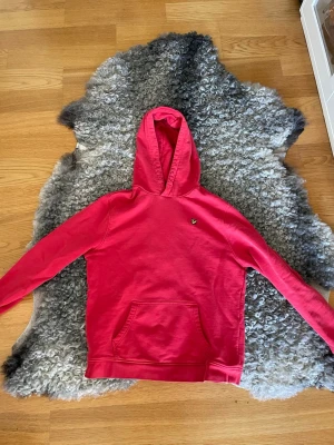 Röd hoodie från Lyle & Scott - Snygg röd hoodie från Lyle & Scott med klassisk huva och magficka. Liten gul logga på bröstet ger en clean look. Tillverkad i mjukt bomullsmaterial som är skönt att ha på sig. Perfekt för dig som vill ha en enkel men stilren hoodie.