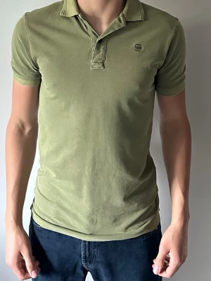 Olivgrön piké från G-Star RAW - Snygg olivgrön pikétröja från G-Star RAW med klassisk krage och korta ärmar. Tröjan har diskret logga på bröstet och är tillverkad i mjuk bomull för en bekväm känsla. Passformen är normal och stilen är enkel men stilren.