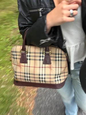 Burberry rutig handväska i läder - Klassisk Burberry handväska med beige, rött och svart rutmönster. JÄTTESNYGG burberry väska köpt från sellpy. En defekt på kanten av ena sidan, visas på sista bilden. Inte något som syns tydligt eller i alla ljus. Kan hitta äkthetsbevis vid intresse. Finns även nyckel till låset så man kan låsa dragkedjan på väskan🤗