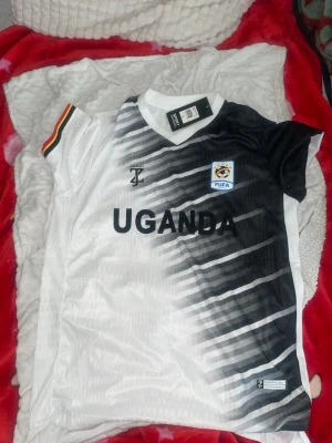 Uganda FUFA matchtröja svart/vit - Snygg Uganda FUFA matchtröja från Janzi i svart och vitt med diagonal design och trycket 'UGANDA' på bröstet. Kort ärm med röd, gul och svart detalj på ena ärmen. Tillverkad i lätt och ventilerande material, perfekt för fotboll eller träning.