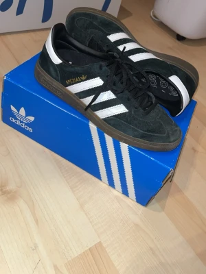 Adidas Spezial svarta sneakers - Snygga Adidas Spezial sneakers i svart mocka med klassiska vita ränder på sidorna och guldfärgad text. Skorna har en brun gummisula och svart snörning. Insidan har Adidas-logga och hälen är vit. Perfekta för dig som gillar retrostil och streetwear.