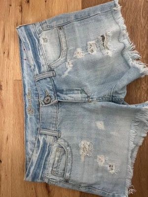 Ljusblå slitna jeansshorts från Guess - Säljer ett par ljusblå jeansshorts från Guess med slitna detaljer och fransig kant. Shortsen har klassisk femficksdesign, knappgylf och logga bak på linningen. Perfekta för sommaren och ger en avslappnad vibe.