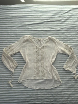 Billabong vit boho-blus med broderi  - Säljer en vit boho-inspirerad blus från Billabong med V-ringning och broderade detaljer på framsidan och ärmarna. Blusen har långa ärmar med knytband och tofsar vid ärmsluten. Tillverkad i ett lätt och luftigt bomullstyg som är lite tjockare och håller dig varm när du behöver det. 