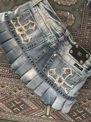 Ljusblå jeanskjol från Miss Me - Säljer en kort ljusblå jeanskjol från Miss Me med plisserad nederkant och dekorativa bakfickor med korsdetaljer och nitar. Kjolen har en cool streetstil och passar perfekt till sneakers eller boots.