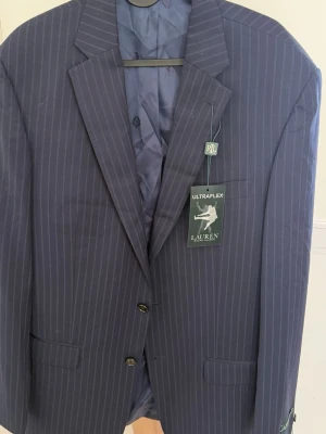 Randig blå kavaj från Lauren Ralph Lauren - Snygg marinblå kavaj från Lauren Ralph Lauren med tunna vita ränder. Klassisk passform med två knappar framtill, fickor och elegant foder. Perfekt för dig som vill ha en stilren och tidlös look. Modellen heter Lexington och materialet är polyester.