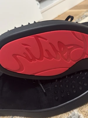 Svarta Louboutin sneakers med nitar - Snygga svarta sneakers från Christian Louboutin med klassisk röd sula och svarta nitar på tån. Skorna har snörning, är låga och tillverkade i mocka med läderdetaljer. Perfekta för dig som vill sticka ut med en edgy och exklusiv look.