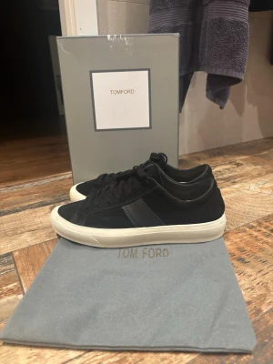 Svarta Tom Ford Skor - Snygga svarta sneakers från Tom Ford i mocka med svarta skosnören och vit sula. Klassisk låg modell med diskret design och en svart läderdetalj på sidan. Perfekta för dig som gillar stilrena och exklusiva sneakers. Dustbag och boxen tillkommer självklart! Skriv vid funderingar.