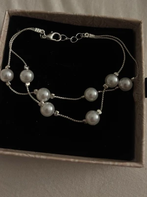 Dubbelradigt armband med pärlor - Snyggt armband med två tunna silverfärgade kedjor och vita pärlor jämnt fördelade längs banden. Armbandet har en klassisk karbinhake och en elegant, stilren design som passar perfekt till din vardagsstil. Perfekt accessoar för att lyfta vilken outfit som helst.