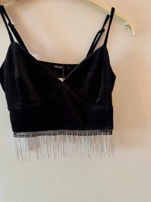 Svart topp BikBok - Snygg svart croppad topp topp från BikBok i mjuk sammet. Toppen har tunna axelband, v-ringning och glittriga strassfransar längs nederkanten. Helt ny och oanvänd. Perfekt för festliga tillfällen!