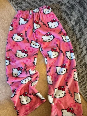 Hello Kitty fleecebyxor loungewear - Mysiga fleecebyxor med Hello Kitty-motiv i två färger: ett par rosa och ett par svarta. Båda har elastisk midja och är täckta av söta Hello Kitty-ansikten och rosetter. Perfekta för chill hemma eller sleepovers. Materialet är mjukt och värmande fleece.
