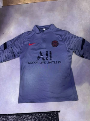 Grå PSG träningströja från Nike - Snygg grå långärmad träningströja från PSG med svart och rött tryck. Halvlång dragkedja framtill, PSG-logga på bröstet och texten 'Accor Live Limitless'. Mönstrade ärmar och 'Visit Rwanda' på ryggen. Perfekt för fotbollsträning.