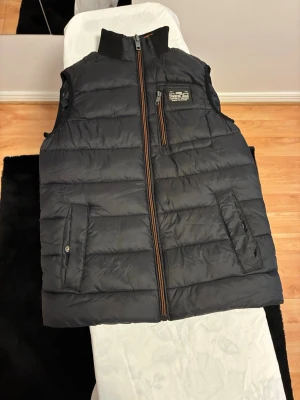 Svart dunväst från Petrol Industries - Snygg svart dunväst från Petrol Industries med hög krage och orange detaljer på dragkedjan. Västen har två framfickor med knapp och en bröstficka med dragkedja samt en patch med logga på bröstet. Perfekt för lager-på-lager och streetwear.