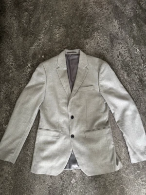 Ljusgrå kavaj från Riley Slim Fit - Stilren ljusgrå kavaj från Riley i slim fit-modell. Kavajen har två knappar framtill, två fickor med lock och en bröstficka. Fodrad insida i grått tyg och klassisk krage. Perfekt för en dressad look.
