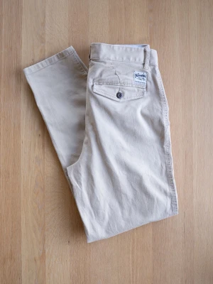 Beige chinos från The Stronghold - Snygga beige chinos från The Stronghold med relaxed tapered passform. Klassisk design med bälteshällor, knappgylf och två sidofickor. Tillverkade i ett mjukt och slitstarkt bomullstyg som ger en clean och stilren look.