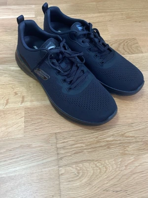Svarta Skechers sneakers i mesh - Svarta sneakers från Skechers med sportig design och ventilerande meshmaterial. Skorna har snörning, diskret logga på sidan och en mjuk, stötdämpande sula för extra komfort. Perfekta för dig som gillar en clean och stilren look.