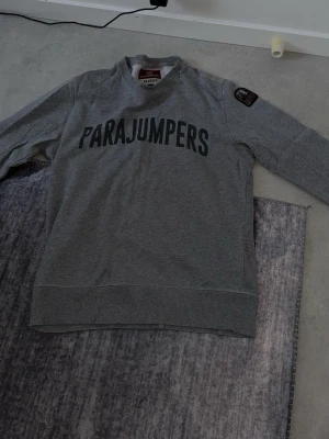 Grå sweatshirt från Parajumpers - Snygg grå sweatshirt från Parajumpers med stort tryck på bröstet och patch på ärmen. Tröjan har rund halsringning, ribbade muddar och är tillverkad i mjukt bomullsmaterial. Perfekt för en avslappnad och trendig look.