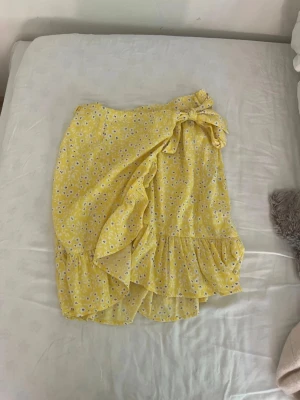 Gul blommig omlottkjol med volang - Söt gul omlottkjol med små vita blommor och volangkant. Kjolen knyts i midjan med en rosett och har en luftig, somrig känsla. Det är i storlek xs och den är ifrån Only
