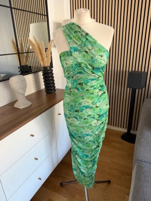 Grön one shoulder Adoore klänning strl 36 - Snygg grön långklänning med blommigt mönster och one shoulder-design. Klänningen har draperade detaljer och smickrande passform som framhäver figuren. Perfekt för dig som vill sticka ut med en unik och trendig look.
